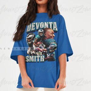 May include: A blue t-shirt featuring a graphic of football player DeVonta Smith. The design includes the player's name in distressed text and multiple images of him in action. The shirt is worn by a person.
