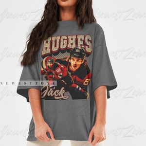 May include: A gray oversized t-shirt with a vintage-style graphic of a hockey player wearing a red and white jersey with the name "Hughes" and the number "86" on the back. The text "Jack" is printed below the graphic.