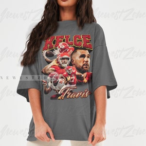 May include: Gray oversized t-shirt with a vintage-style graphic of a football player wearing a red jersey with the name "Kelce" and the number "87" printed on it. The text "Travis" is printed below the graphic.