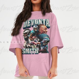 May include: A light pink oversized t-shirt featuring a graphic of football player DeVonta Smith. The design includes the player's name and images of him in action, with the team's colors and logo.