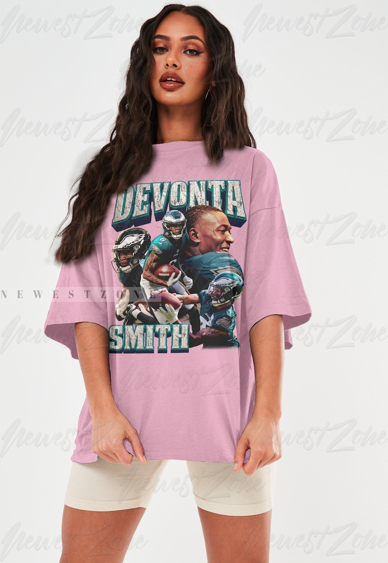 May include: A light pink oversized t-shirt featuring a graphic of football player DeVonta Smith. The design includes the player's name and images of him in action, with the team's colors and logo.