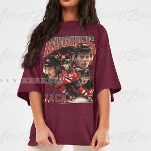 Puede incluir: Una camiseta de gran tama&ntilde;o de color granate con un gr&aacute;fico de estilo vintage de jugadores de hockey. El gr&aacute;fico incluye el texto "HUGHES" y "JACK" y el n&uacute;mero "86".