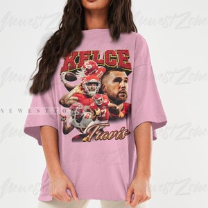 May include: A pink oversized t-shirt with a vintage-style graphic of a football player wearing a red jersey with the name "Kelce" and the number "87" on it. The text "Travis" is printed below the graphic.