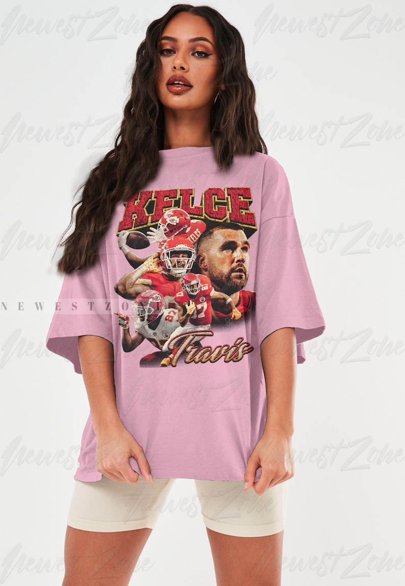 May include: A pink oversized t-shirt with a vintage-style graphic of a football player wearing a red jersey with the name "Kelce" and the number "87" on it. The text "Travis" is printed below the graphic.