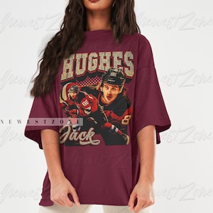 May include: A maroon t-shirt with a vintage-style graphic of a hockey player wearing a red and white jersey with the number 86 on it. The text "HUGHES" and "Jack" are printed on the shirt.