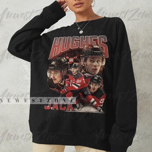 Puede incluir: Sudadera negra con un estampado gr&aacute;fico de un jugador de hockey, Jack Hughes, que lleva una camiseta roja y blanca con el n&uacute;mero 86. El texto "HUGHES" y "JACK" est&aacute; impreso en rojo en la sudadera.