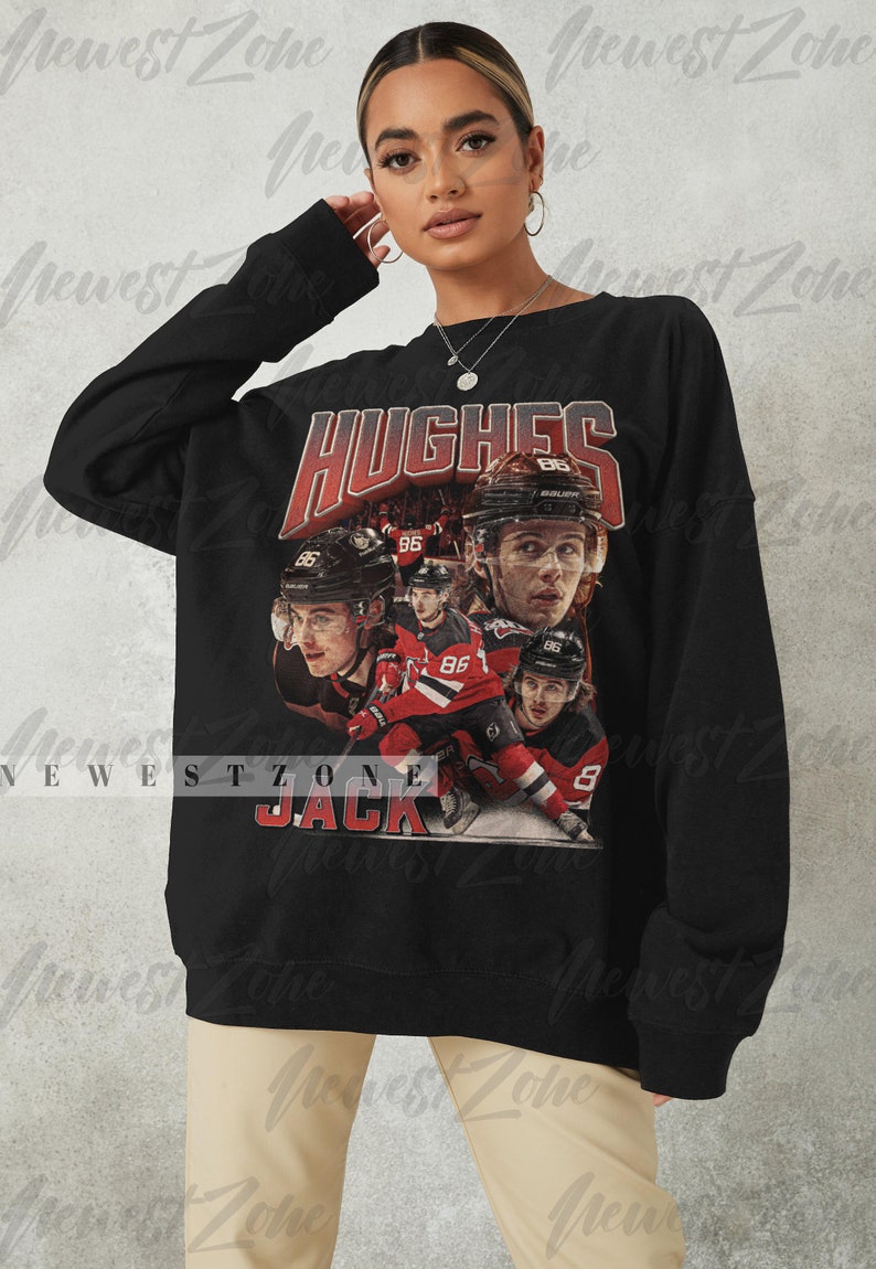 Puede incluir: Sudadera negra con un estampado gr&aacute;fico de un jugador de hockey, Jack Hughes, que lleva una camiseta roja y blanca con el n&uacute;mero 86. El texto "HUGHES" y "JACK" est&aacute; impreso en rojo en la sudadera.