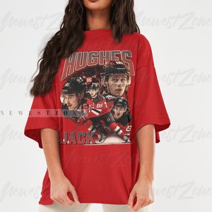 Puede incluir: Camiseta roja de gran tama&ntilde;o con un gr&aacute;fico de estilo vintage de jugadores de hockey que llevan camisetas rojas y blancas. El gr&aacute;fico incluye el texto "HUGHES" y "JACK".