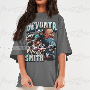 May include: A gray t-shirt featuring a graphic of football player DeVonta Smith. The design includes images of the player in action, with the name "DEVONTA SMITH" in a distressed font. The shirt is a casual, oversized style.