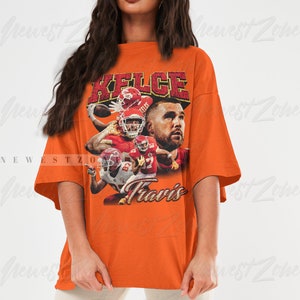 May include: Orange oversized t-shirt with a vintage-style graphic of Travis Kelce, a football player for the Kansas City Chiefs. The graphic features Kelce in action with the text "Kelce" and "Travis" printed on the shirt.