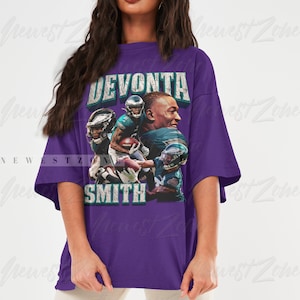 May include: A purple t-shirt featuring a graphic of Devonta Smith, with the text "DEVONTA SMITH" in a distressed font. The graphic includes images of Smith in an Eagles uniform. The shirt is oversized.