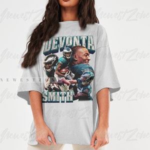 May include: White t-shirt featuring a graphic of football player DeVonta Smith. The design includes the player's name in a distressed font, along with images of him in action. The shirt has short sleeves and a relaxed fit, suitable for casual wear.