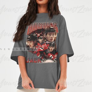 Puede incluir: Una camiseta gris de gran tama&ntilde;o con un gr&aacute;fico de estilo vintage de jugadores de hockey que llevan camisetas rojas y blancas. El gr&aacute;fico incluye el texto "HUGHES" y "JACK".