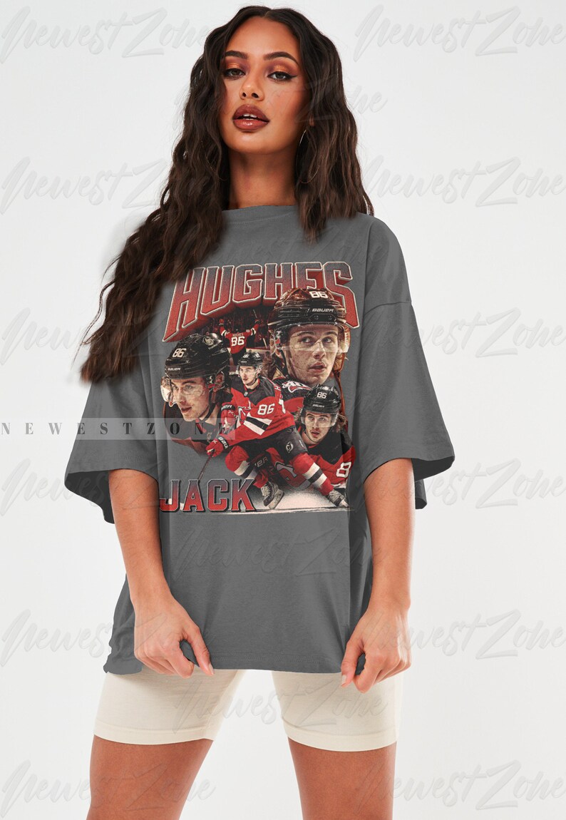 Puede incluir: Una camiseta gris de gran tama&ntilde;o con un gr&aacute;fico de estilo vintage de jugadores de hockey que llevan camisetas rojas y blancas. El gr&aacute;fico incluye el texto "HUGHES" y "JACK".