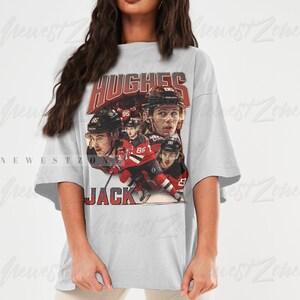 Puede incluir: Una camiseta gris de gran tama&ntilde;o con un gr&aacute;fico de estilo vintage de jugadores de hockey. El gr&aacute;fico presenta el texto "HUGHES" y "JACK" y el n&uacute;mero "86".