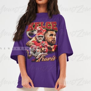 May include: Purple oversized t-shirt with a vintage-style graphic of a football player wearing a red jersey with the name "Kelce" and the number "87" on it. The text "Travis" is printed below the graphic.