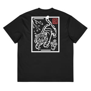 May include: Black t-shirt with a white graphic of a tiger, flowers, and a branch design. The tiger is within a white rectangular frame. A red square with Asian characters is in the upper right corner.