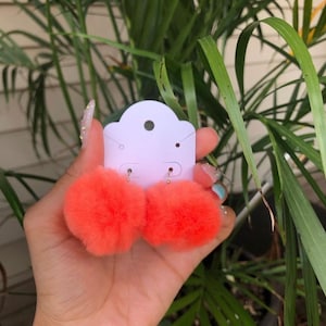 以下が含まれることがあります： 鮮やかなオレンジ色のふわふわポンポンピアス。ピアスは、上部に小さな穴が開いた白いピアスディスプレイカードに取り付けられています。背景には緑の葉があります。