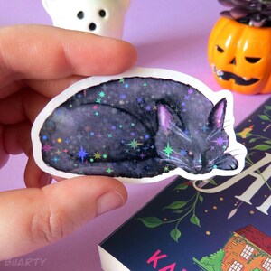 Peut inclure: Autocollant représentant un chat noir endormi, orné d'étoiles holographiques. Le chat est enroulé sur lui-même, avec des détails violets sur les oreilles. Un petit fantôme et une citrouille sont en arrière-plan.
