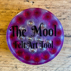 Op de afbeelding: Een rond vilten gereedschap in paars en wit met de tekst "The Mool Felt Art Tool" en de website "www.moymackaygallery.com".