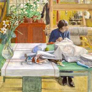 May include: A woman sits at a table sewing a white garment. The table is covered with a white tablecloth and a green runner. There are several pieces of fabric and a vase of white flowers on the table. The woman is wearing a blue sweater and has her hair pulled back. The room is brightly lit and there is a loom in the background.