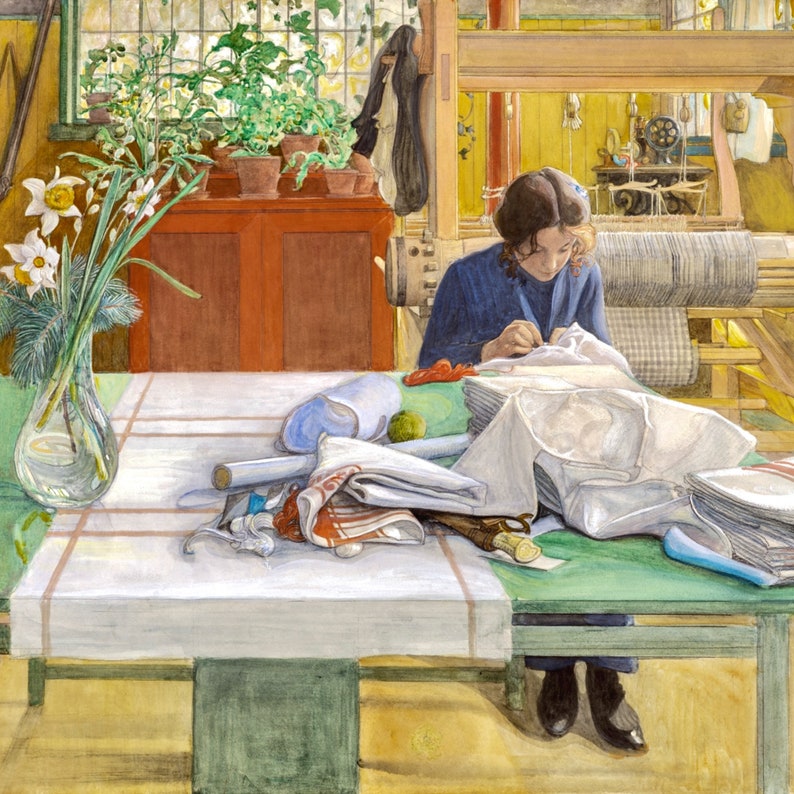 May include: A woman sits at a table sewing a white garment. The table is covered with a white tablecloth and a green runner. There are several pieces of fabric and a vase of white flowers on the table. The woman is wearing a blue sweater and has her hair pulled back. The room is brightly lit and there is a loom in the background.