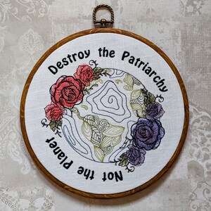 May include: Embroidered hoop art with a white background and a colorful floral design. The text "Destroy the Patriarchy Not the Planet" is embroidered in black.