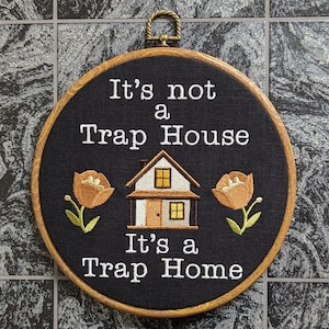May include: Embroidered hoop art with a black background and white text that reads "It's not a Trap House It's a Trap Home". The hoop also features a small white house with orange windows and two orange flowers.