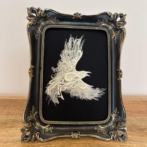 May include: An embroidered artwork of a bird in flight, set against a black backdrop within a decorative black frame with gold detailing. The bird is cream-colored, with its wings outstretched and beak open.