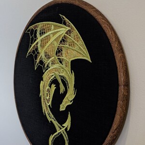 Puede incluir: Un dise&ntilde;o de drag&oacute;n bordado en oro sobre tela negra en un aro de bordado de madera.