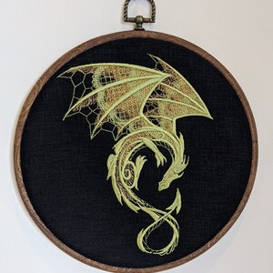 Puede incluir: Dise&ntilde;o de drag&oacute;n verde bordado sobre un lienzo circular negro, enmarcado en un aro de madera oscura.