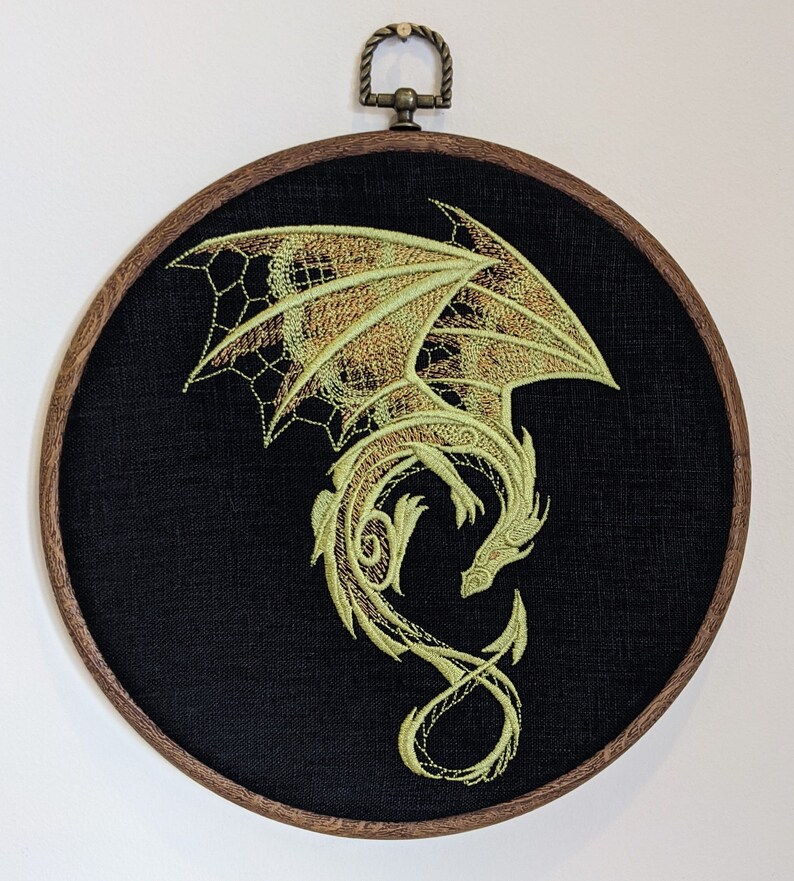 Puede incluir: Dise&ntilde;o de drag&oacute;n verde bordado sobre un lienzo circular negro, enmarcado en un aro de madera oscura.