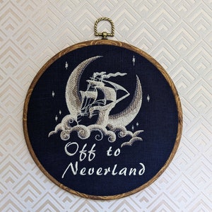 May include: A dark blue round embroidery hoop with a white embroidered design of a ship sailing on a crescent moon with clouds. The text "Off to Neverland" is embroidered below the ship.