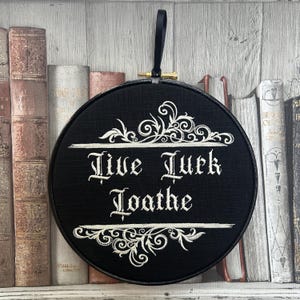 May include: A circular black embroidery hoop featuring white stitched text that reads Live Lurk Loathe, accented with decorative white scrollwork above and below the lettering, and a black ribbon loop for hanging at the top.