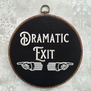 May include: A black and white embroidered sign with a wooden frame. The sign reads "Dramatic Exit" with two white embroidered pointing hands.