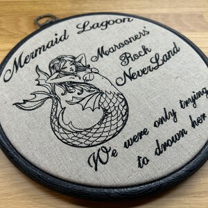 May include: A circular embroidery hoop containing beige fabric with black thread embroidery. The design features a mermaid illustration and the text: Mermaid Lagoon, Marooner's Rock, NeverLand, We were only trying to drown her. The hoop has a black frame.