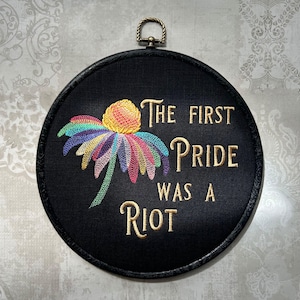 May include: A black, round embroidery hoop with the text "The First Pride Was A Riot" in gold thread. A colorful embroidered flower with rainbow petals is centered on the black fabric. The hoop has a brass-colored hanging loop.