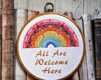 Puede incluir: Arte de bordado en aro con un diseño de arcoíris y el texto "All Are Welcome Here" en fuente blanca. El aro está rodeado de una colección de libros.