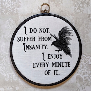 May include: Black and white embroidered hoop art featuring a raven and the text "I do not suffer from insanity. I enjoy every minute of it."