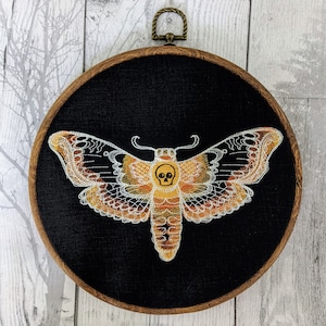 Peut inclure: Un cerceau de broderie noir avec un papillon de nuit jaune et orange avec un motif de crâne sur son corps. Le papillon de nuit est brodé sur un tissu noir.
