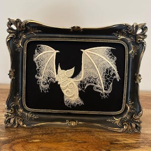 May include: An embroidered bat design in cream thread on black fabric, presented in an ornate black frame with gold detailing. The bat's wings are spread, showcasing delicate lace-like patterns. The frame's design adds a touch of vintage elegance.