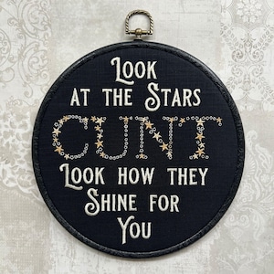 May include: A circular black embroidery hoop wall hanging featuring white stitched text that reads Look at the stars, followed by the word CUNT spelled out in gold star-shaped sequins, and the text Look how they shine for you below.