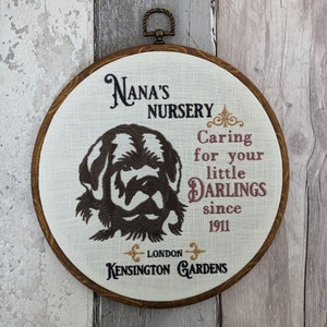 May include: A brown wooden embroidery hoop with white fabric inside. The fabric has an embroidered image of a brown dog with the text "Nana's Nursery Caring for your little Darlings since 1911 London Kensington Gardens".