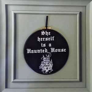 May include: Black embroidery hoop with white text that reads "She herself is a Haunted House" and a white illustration of a haunted house with a crescent moon above it.