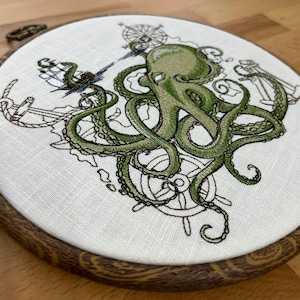 May include: Embroidered hoop art featuring a green octopus with tentacles wrapped around a ship's wheel and anchor. The design is on a white linen background.