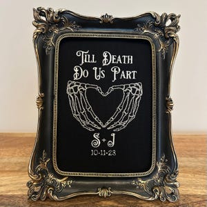 May include: An ornate black framed artwork with gold detailing. The black background features white embroidered skeleton hands forming a heart, with the text "Till Death Do Us Part", initials, and the date 10-11-23.