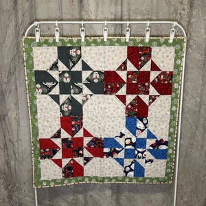 May include: A small, handmade quilt with a patchwork design featuring red, blue, and gray fabric squares with a white background. The quilt is bordered with a green fabric with a white snowflake pattern.