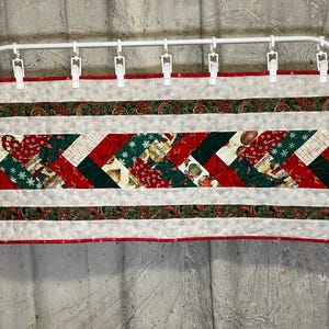 Puede incluir: Una cenefa acolchada con temática navideña con borde rojo y detalles blancos. El diseño central presenta rayas diagonales de telas rojas, verdes y estampadas. Está sujeta a una barra de metal blanca con pinzas.