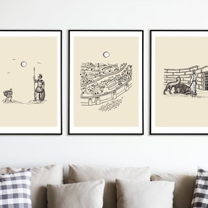 May include: Three black framed prints with beige backgrounds and black ink drawings. The first print shows a person walking away from a soldier holding a spear. The second print shows a city with a river running through it. The third print shows a person standing in front of a cage with a lion inside.