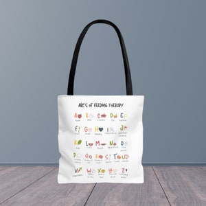 May include: White tote bag with black handles. The bag features a colourful alphabet chart with illustrations and words related to feeding therapy. The text at the top of the chart reads "ABC's of Feeding Therapy".